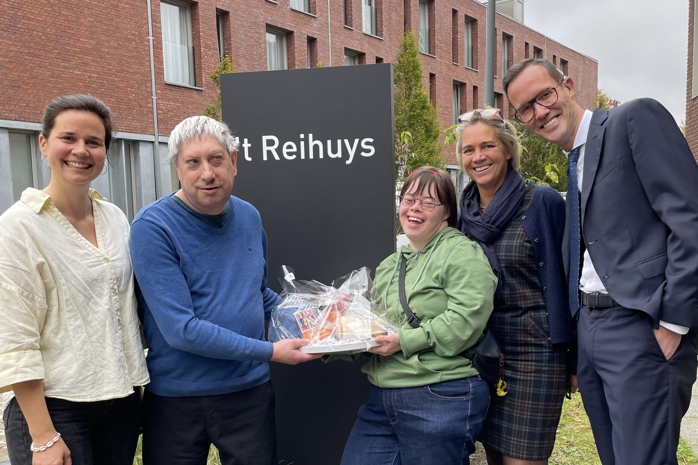 Minister Rob Beenders (rechts op de foto) poseert met de directeur en afdelingsverantwoordelijke van Ons Huis, samen met twee bewoners die een geschenkenmand overhandigen aan de minister.