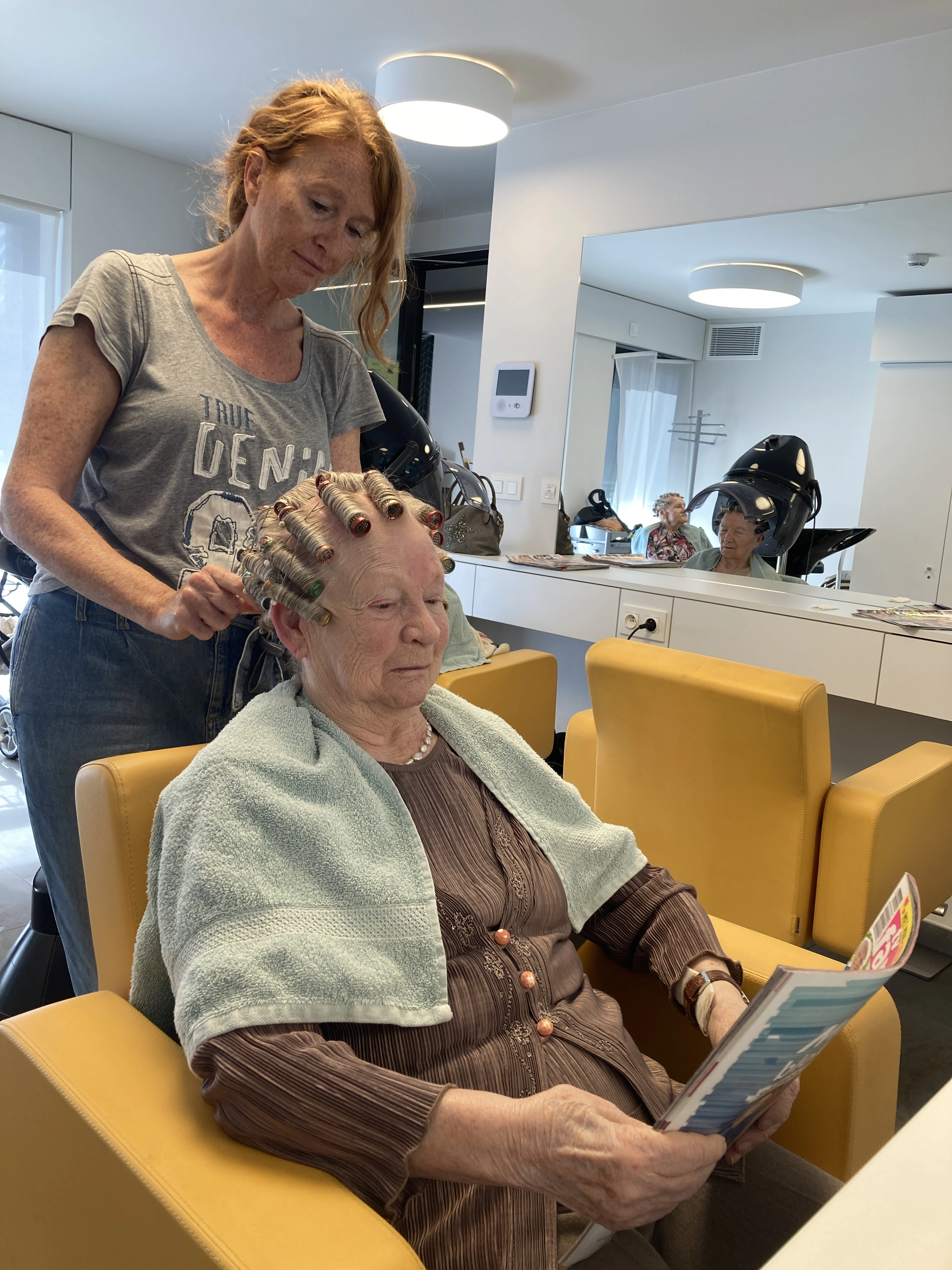 Bewoonster zit in stoel en leest een boekje terwijl de kapper krullen legt